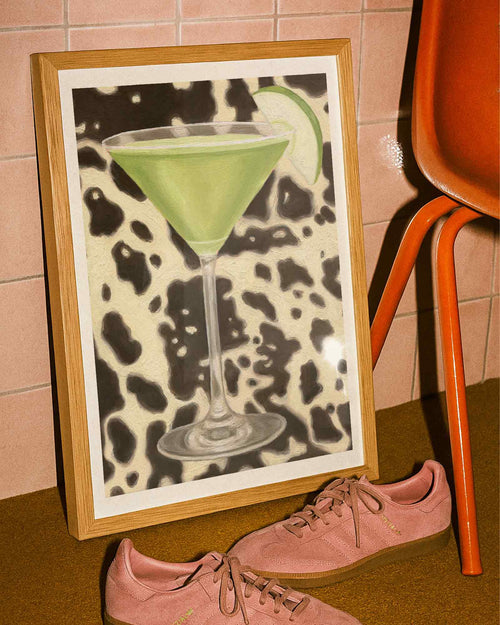 Framed artwork of a green cocktail with a cow print background next to pink sneakers.