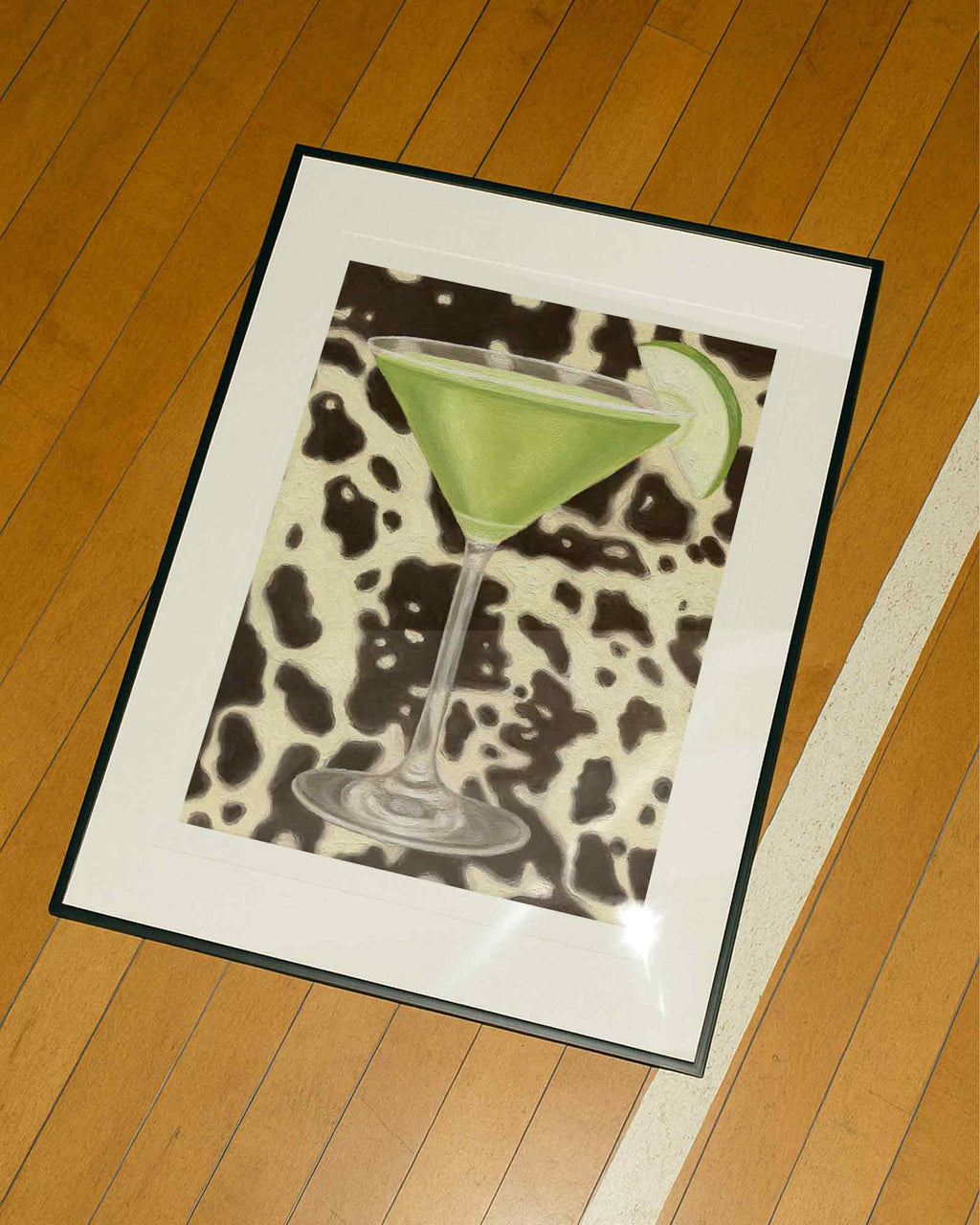 Framed artwork of a green cocktail with a slice of apple on a cow print background, placed on a wooden floor.