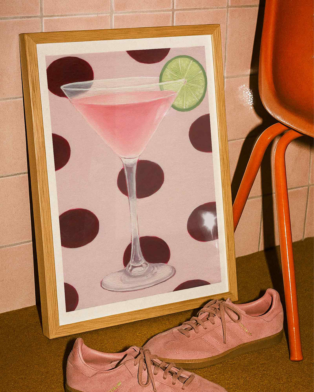 Framed artwork of a pink cocktail with a lime wedge on a polka dot background, placed next to pink sneakers and an orange chair.