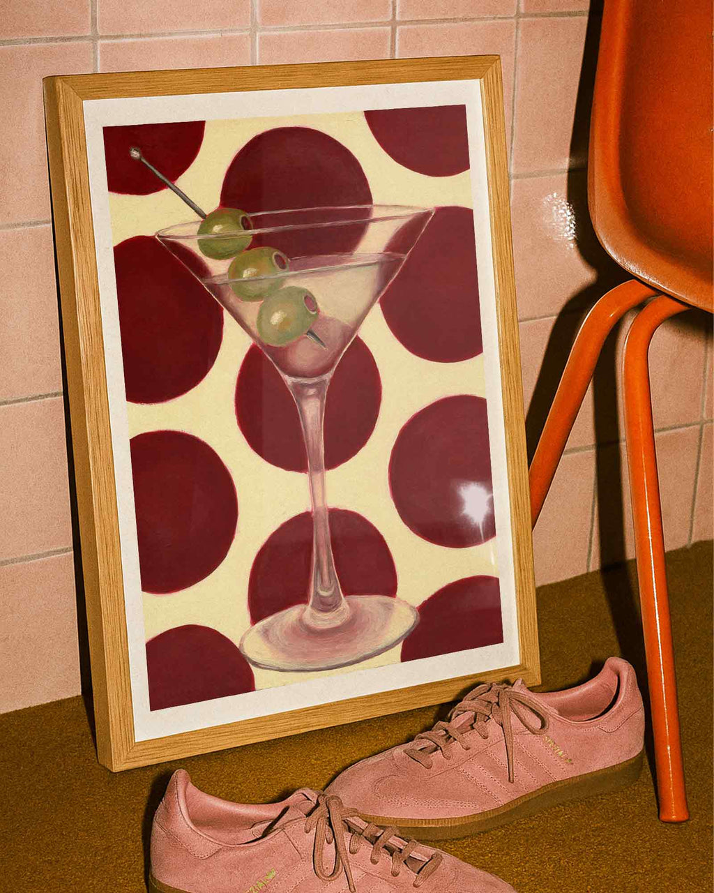 Framed artwork of a martini with olives on a polka dot background, next to pink shoes and an orange chair.