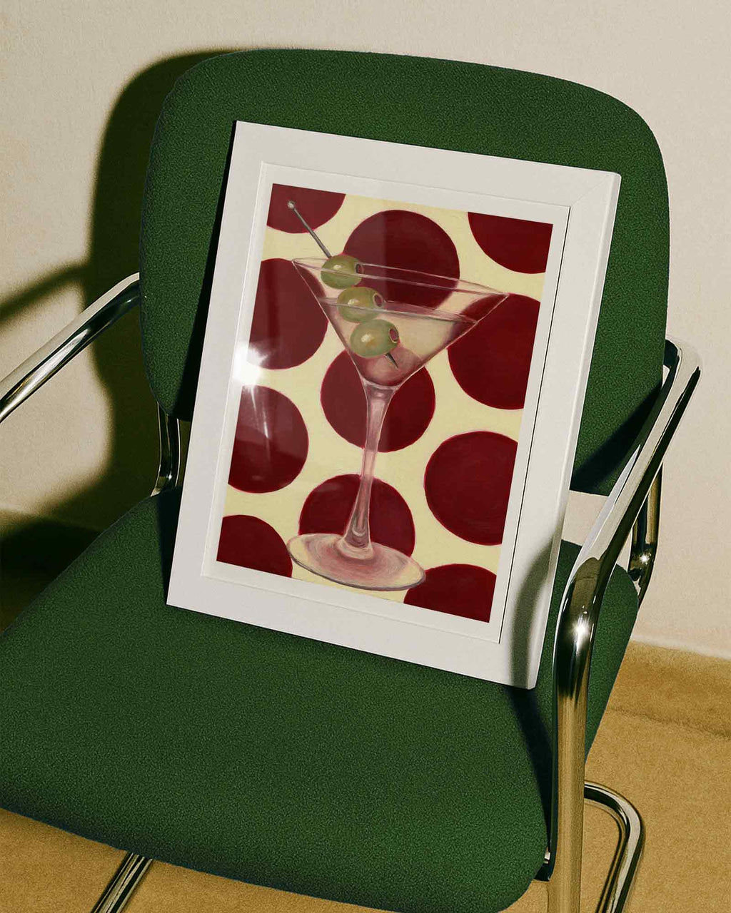 Framed artwork of a martini glass with olives on a red polka dot background, placed on a green chair.