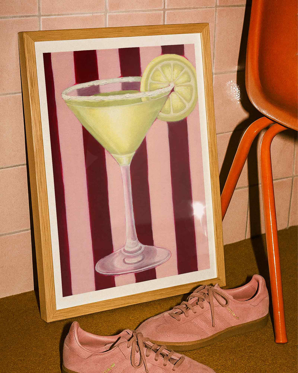 Framed artwork of a cocktail with a lemon slice on a striped background, next to pink shoes and an orange chair.