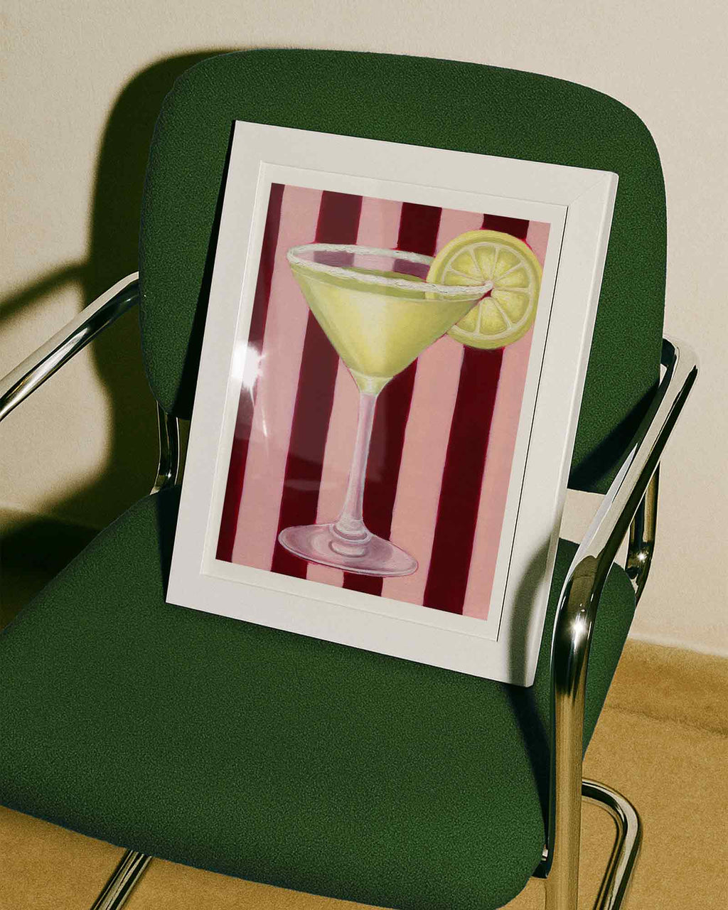 Framed artwork of a cocktail with a lemon wedge on a striped background, placed on a green chair.
