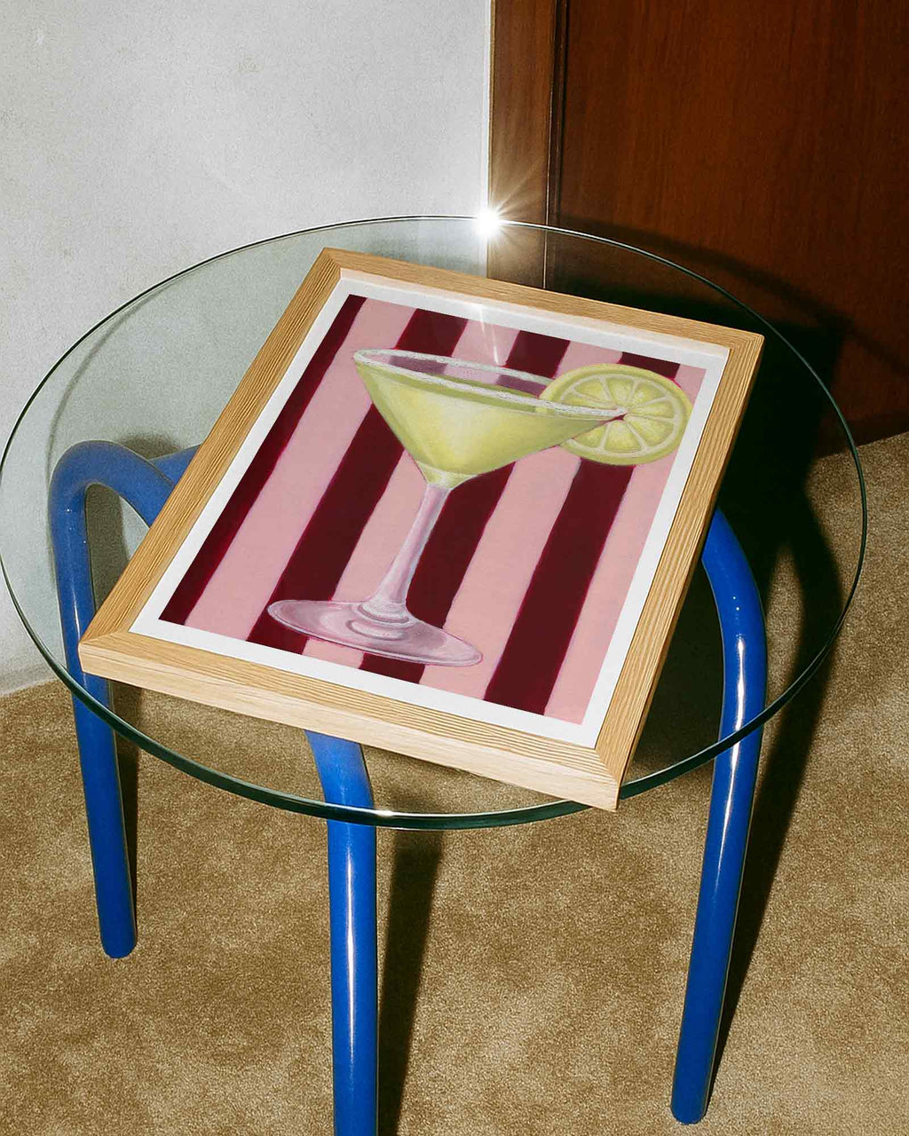 Framed artwork of a cocktail with a lemon wedge on a glass table.
