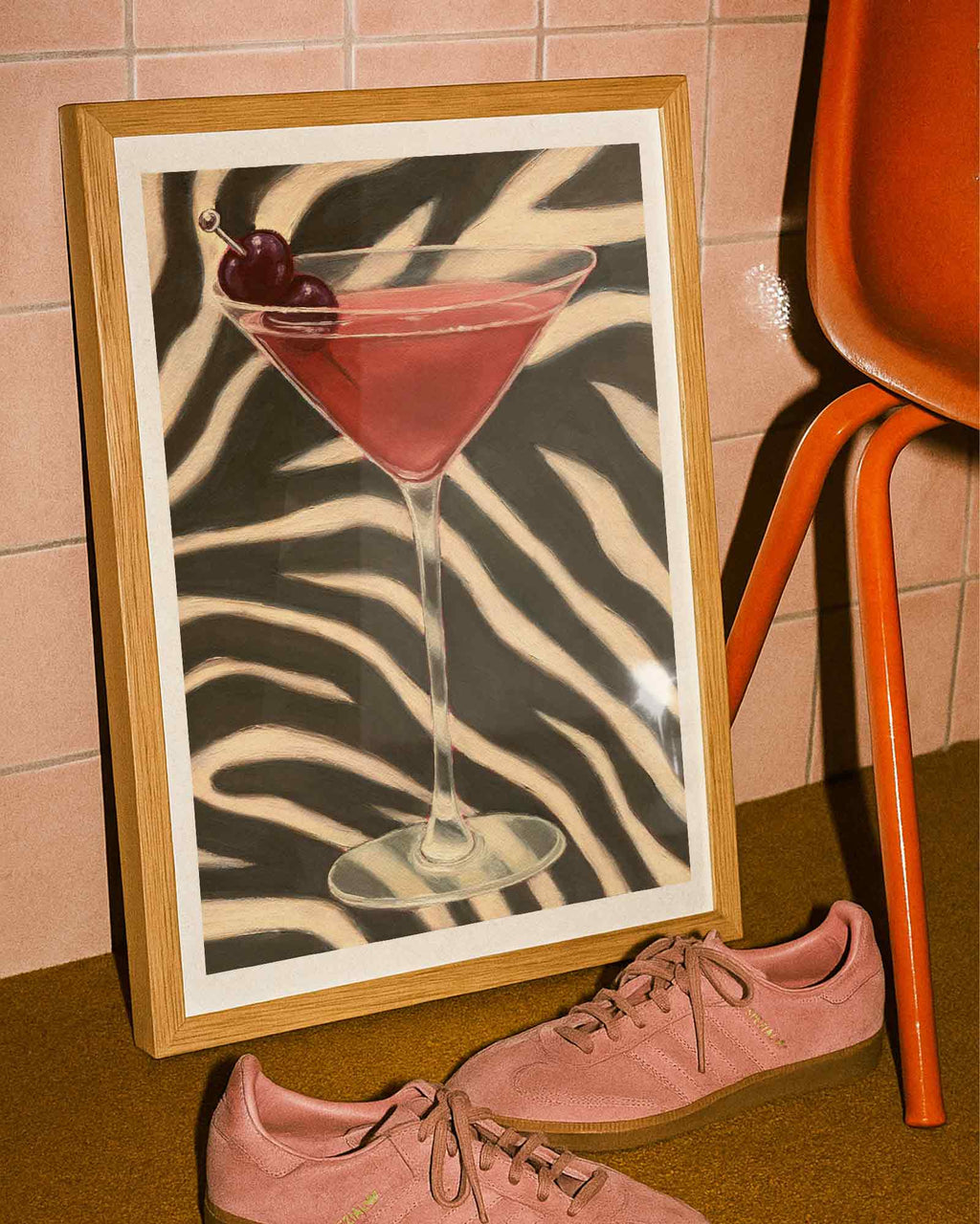 Framed artwork of a cocktail with cherries on a zebra-striped background, next to pink sneakers and an orange chair.
